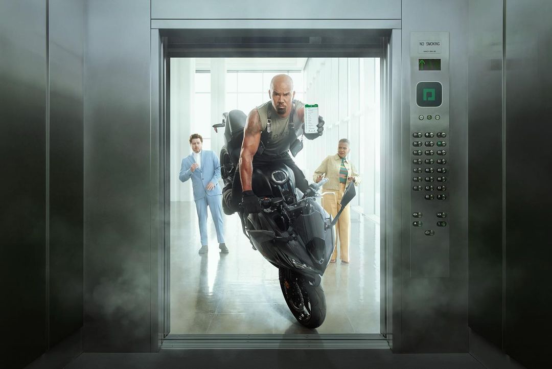 A man rides a motorcycle inside an elevator, holding up a mobile phone, with two other people standing in the elevator's background.